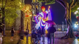 Vista frontal de las Figuras de luz Monumentales de charro y bailarina en el zócalo de Puebla durante las fiestas patrias. De fondo las torres de la Catedral con iluminación dinámica generando photomoments y gente que disfruta en silla de ruedas